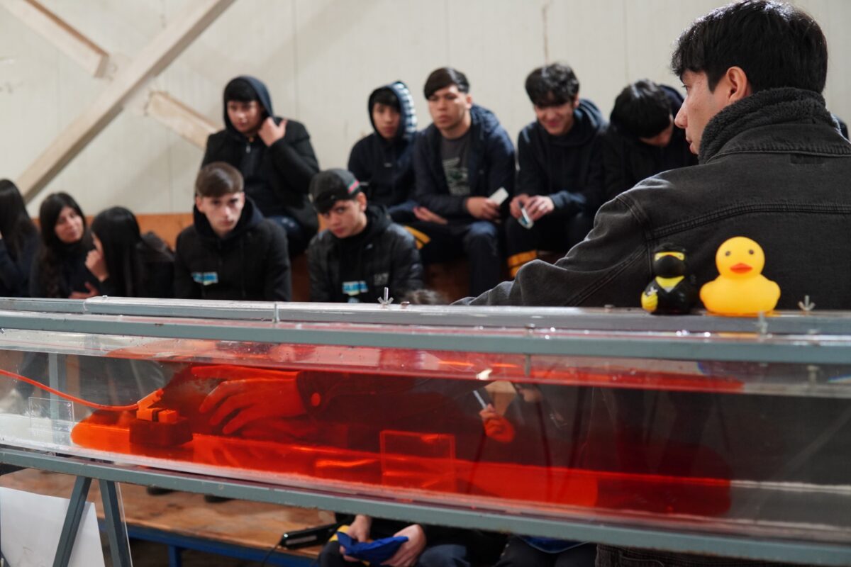 Primer plano de un canal de experimentos con líquido rojo y un objeto sumergido, con estudiantes observando al fondo.
