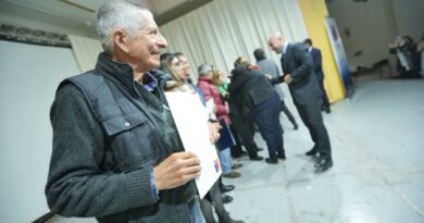 Un hombre mayor sonriente, vistiendo un chaleco oscuro sobre un suéter de lana, sostiene un documento blanco mientras otras personas están de pie en fila detrás de él.