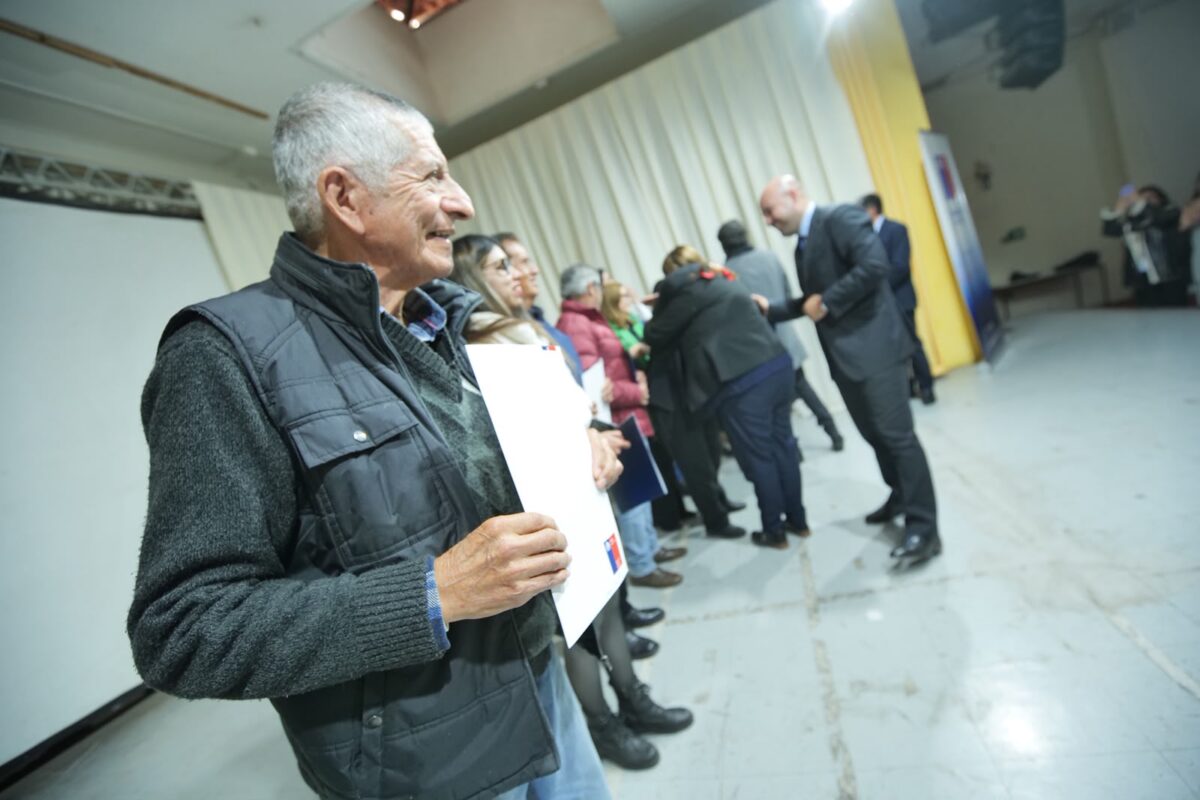 Un hombre mayor sonriente, vistiendo un chaleco oscuro sobre un suéter de lana, sostiene un documento blanco mientras otras personas están de pie en fila detrás de él.