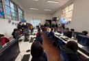 Estudiantes y adultos en un laboratorio de computación con pantallas encendidas y proyectando una imagen en la pared.