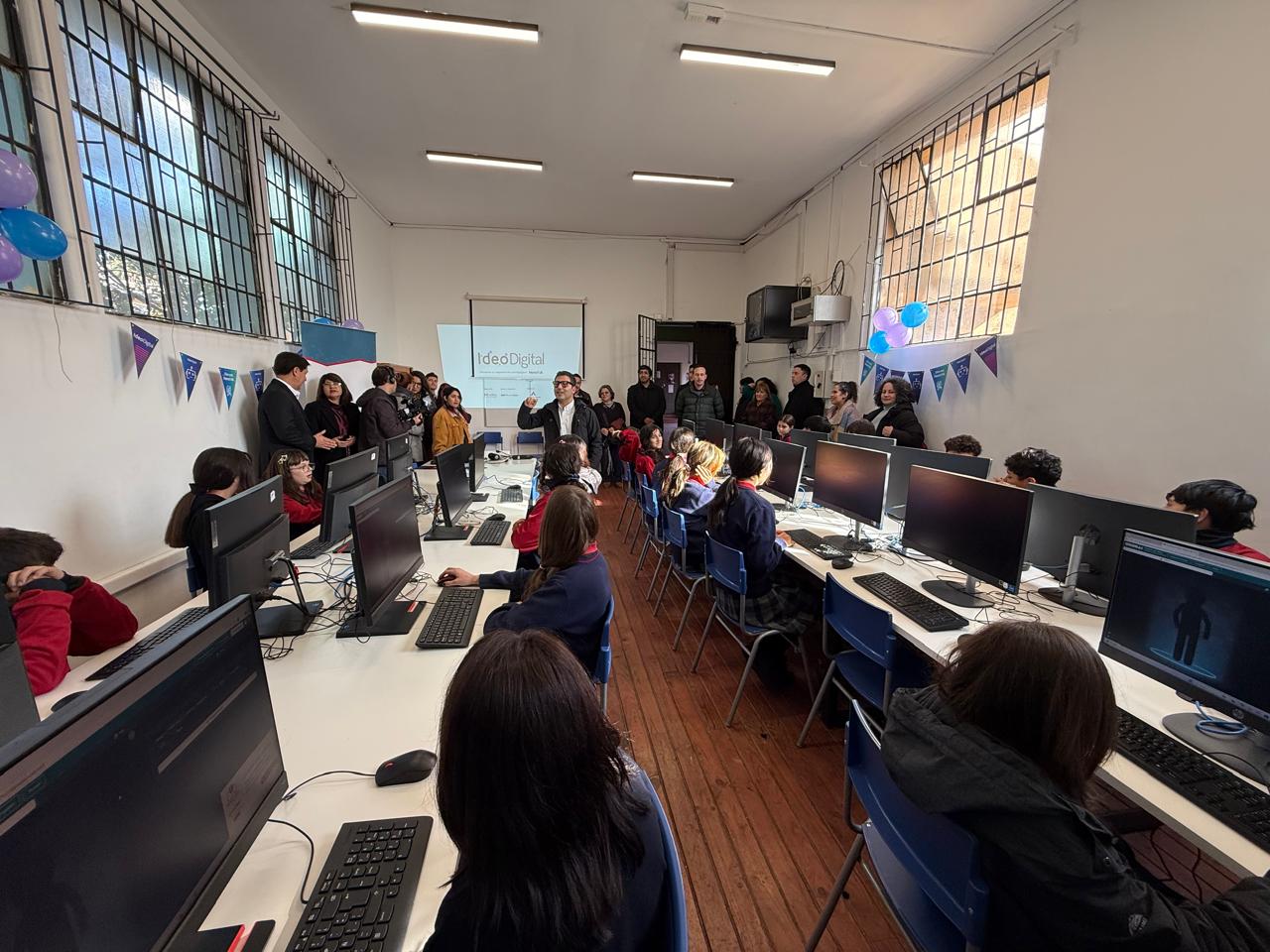 Estudiantes y adultos en un laboratorio de computación con pantallas encendidas y proyectando una imagen en la pared.