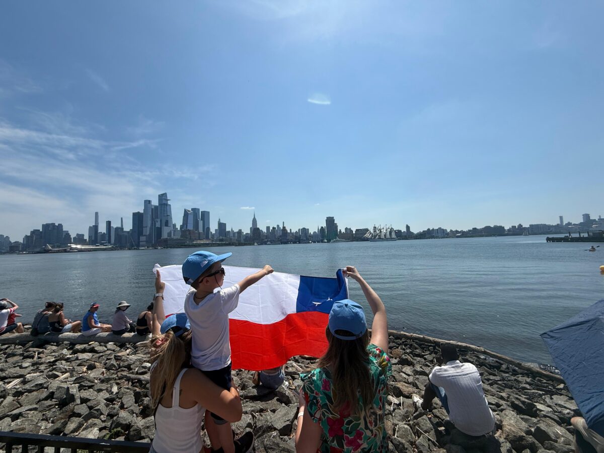 Personas sosteniendo una bandera chilena frente a la bahía de Nueva York con el buque al fondo.