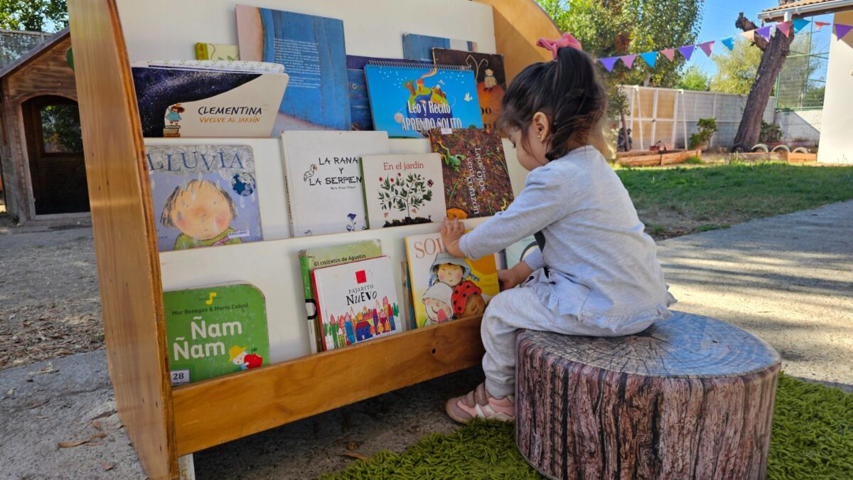 Niña pequeña seleccionando un libro en una biblioteca infantil al aire libre.